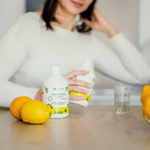 Femme tenant une bouteille de Probiotique Encapsulé Vitamoon avec vitamine C sur une table, accompagnée de citrons frais et d’un verre d’eau, illustrant la consommation quotidienne pour la santé digestive.
